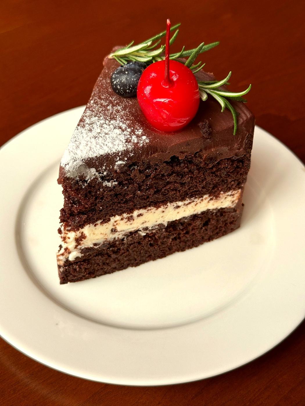 A slice of Matilda cake lifted from the whole: deep chocolate sponge, cream cheese buttercream, cherry and rosemary garnish